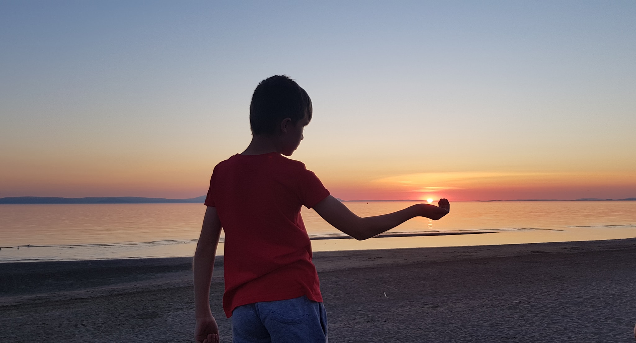 Picture of a boy on a beack at sunset, cupping the sun in his hand