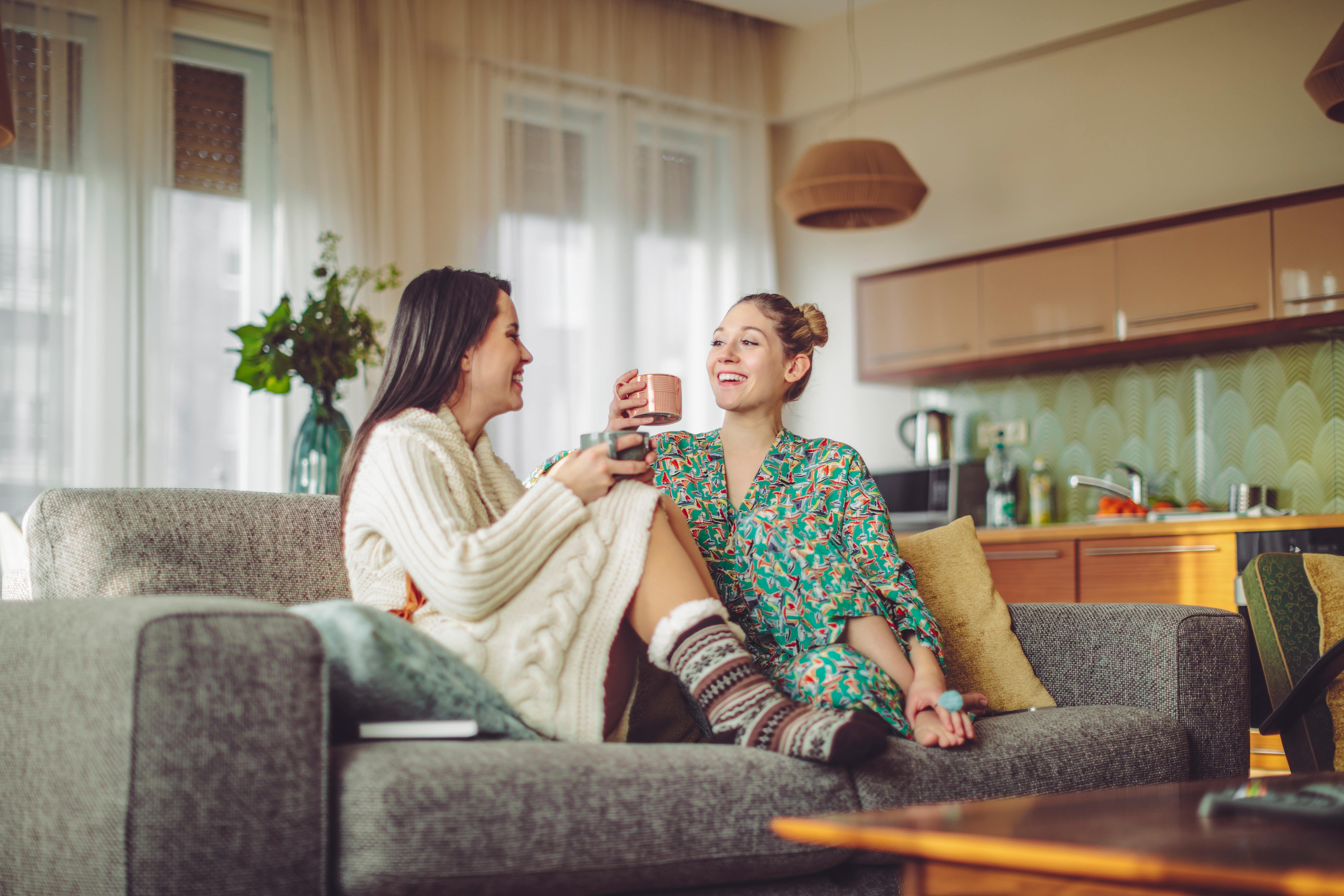 Two women sitting on a grey sofa in a bright living room holding mugs