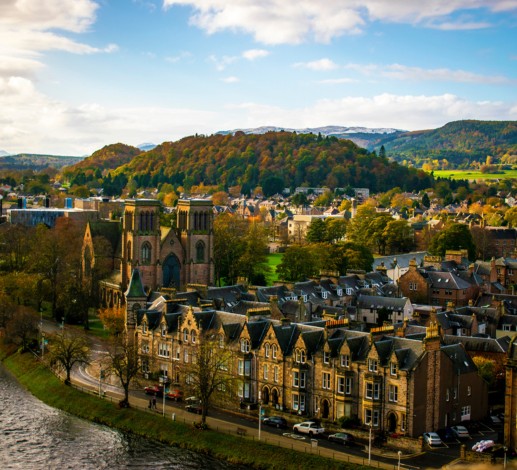 A view of buildings by a river in Inverness, hills thick with trees in autumnal colours are visible beyond the buildings, as well as grass lit by sunlight. The sky is mostly clear, aside from a few white clouds. 