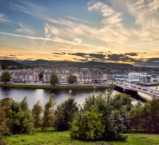 A panoramic view of the city of Inverness. The sky is a mix of blue and an orange hue from the sunsetting.  