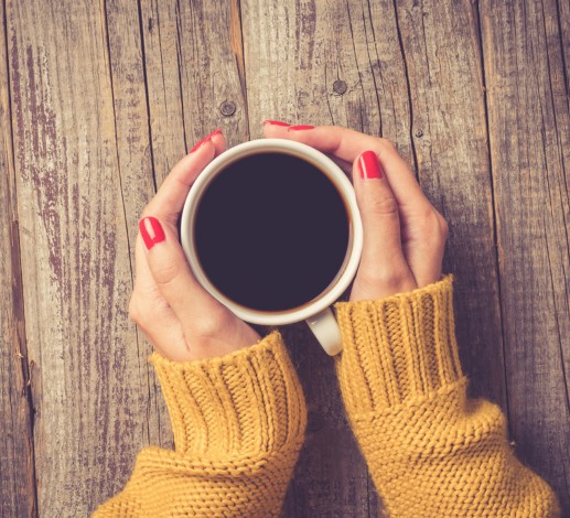 A person is holding a mug of coffee. The sleeves of their yellow jumper are visible on the wooden grain table. 