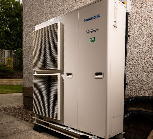 A heat pump installed at the side of a home. A patio style path is visible which leads to a back garden.  
