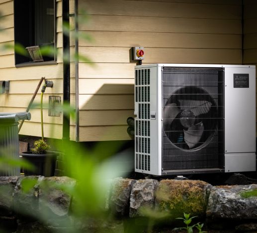 A heat pump installed outside a home. The home has a cream coloured wooden plank exterior.  