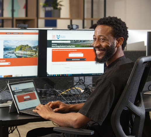 An advisor is smiling and is sitting in front of a laptop and multiple screens displaying the Home Energy Scotland website.