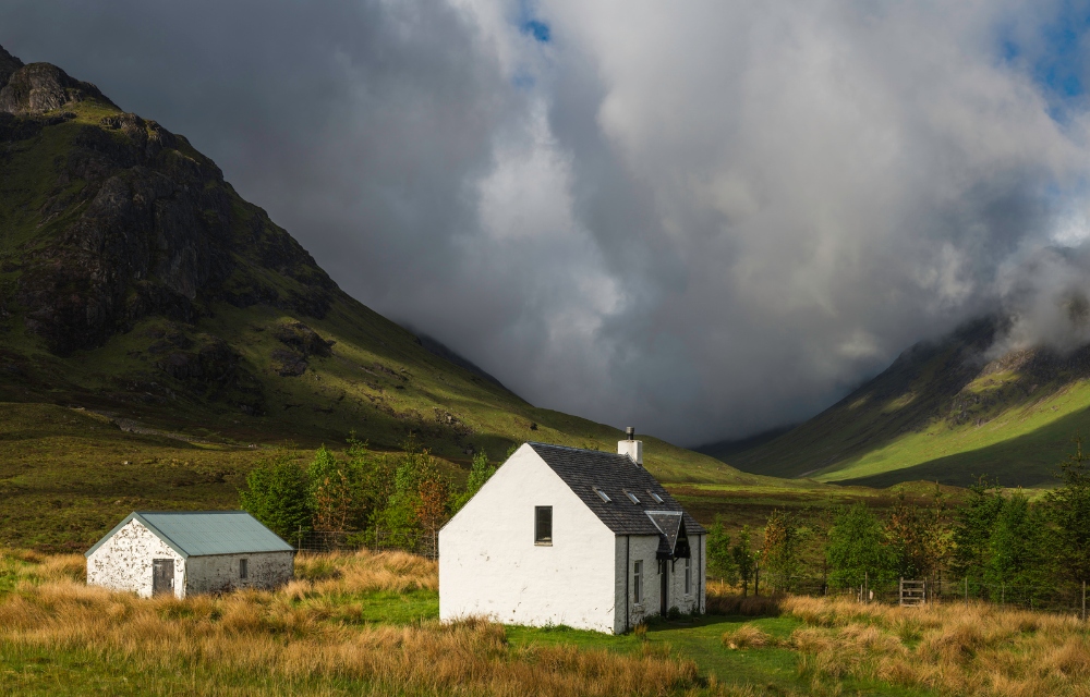 A traditional white-washed crofter's cottage in idyllic Highland glen below dramatic cloudscape sweeping over wild mountain peaks