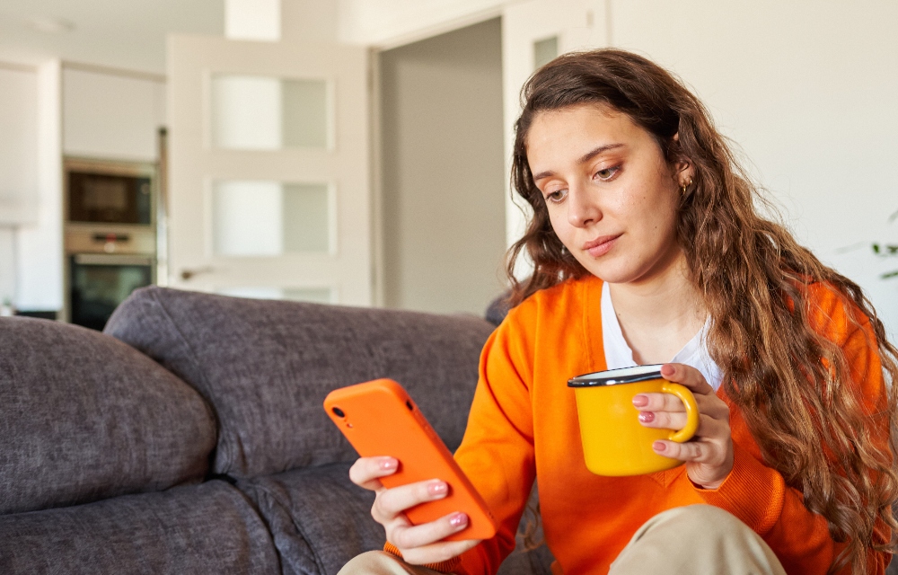 A person wearing an orange jumper is sitting on a couch and is looking at their mobile phone, they are also holding a mug.