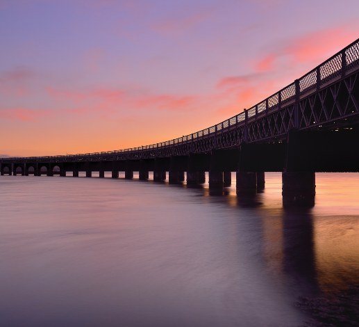 The Tay Rail Bridge at sunset, silhouetted against a pink and purple sky above calm water.