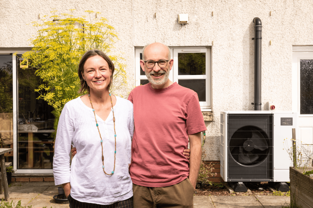 John and Laura standing beside each other in their garden with a heat pump