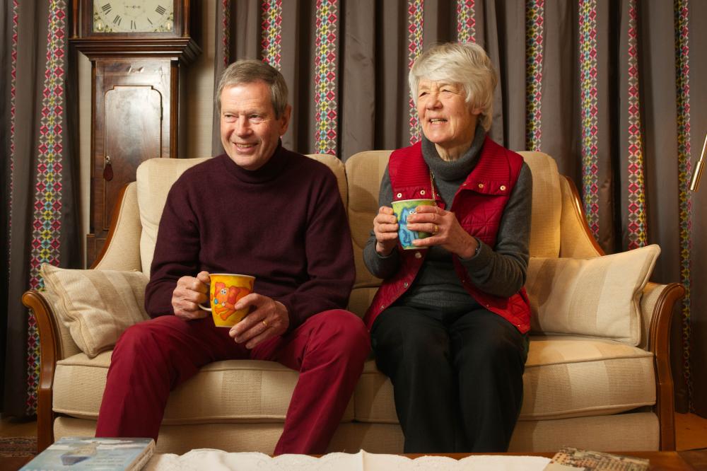 An old couple sitting on their cream sofa next to their grandfather clock. Both of them are holding colourful mugs with cartoon drawings on them