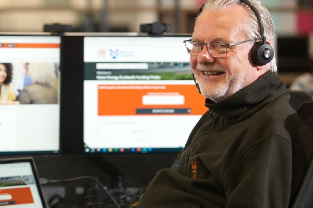 Harry, a Home Energy Scotland advisor, is at a desk and computer wearing a telephone headset, he is smiling