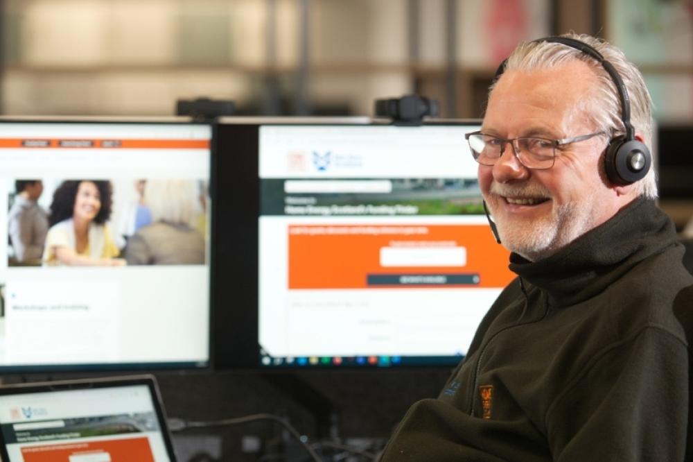 An advisor wearing a headset and Home Energy Scotland branded jacket sits at a laptop, he is smiling
