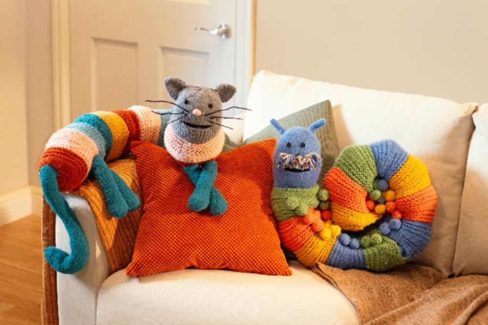 A colourful knitted cat draught excluder is perched on the arm rest of a cream-coloured couch, beside an equally colourful caterpillar  draught excluder. 