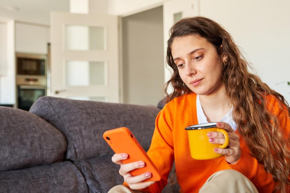 A person wearing an orange jumper is sitting on a couch and is looking at their mobile phone, they are also holding a mug.