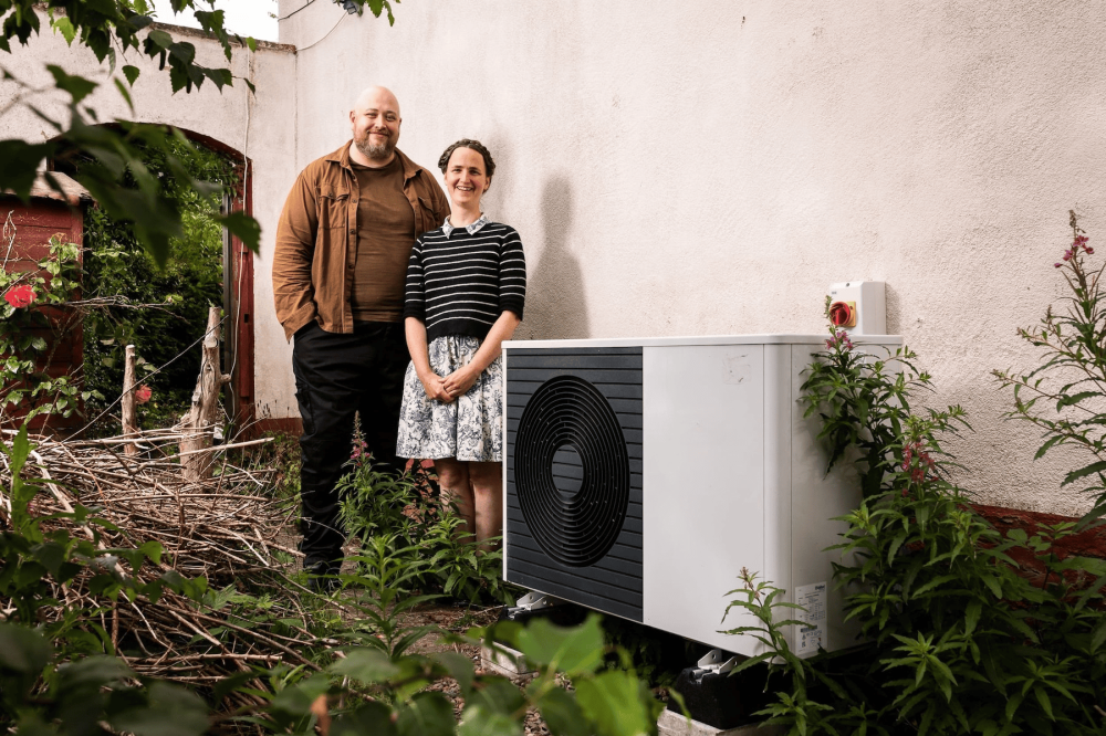 Heid and her partner smiling at the camera while standing beside their air source heat pump