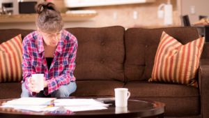 Senior adult woman worries over her past due bills while sitting on home living room sofa.  A pile of invoices on coffee table in front of her.  She is calculating expenses versus budget income.  Frustration among middle-class people.   Great imagery for election season:  home finances, recession, past due bills, mortgage, debt, stress, worry, taxes.